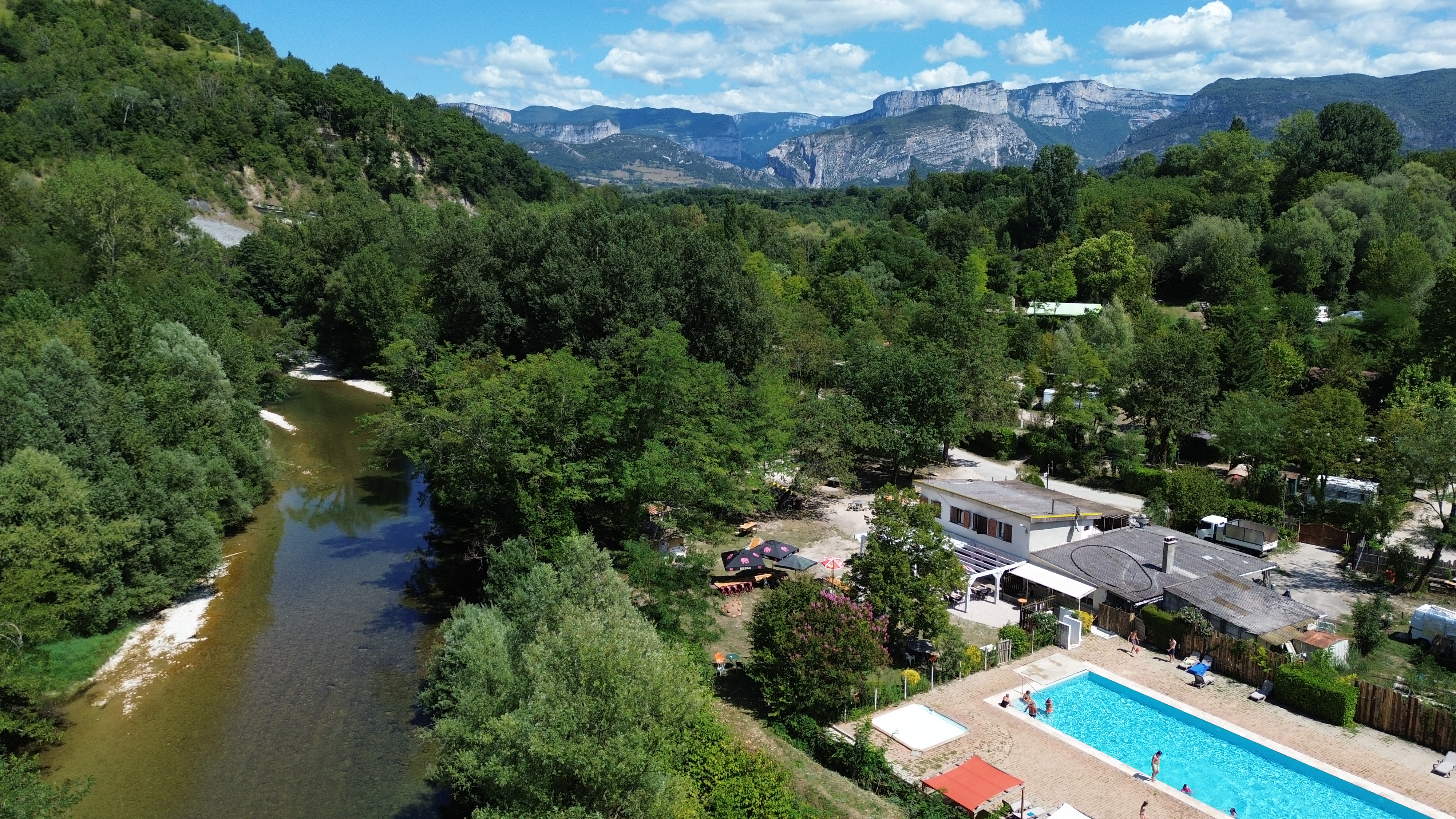Vue aérienne du camping installé au bord d'une rivière, avec sa piscine, ses espaces arborés et les reliefs du Vercors en [...]